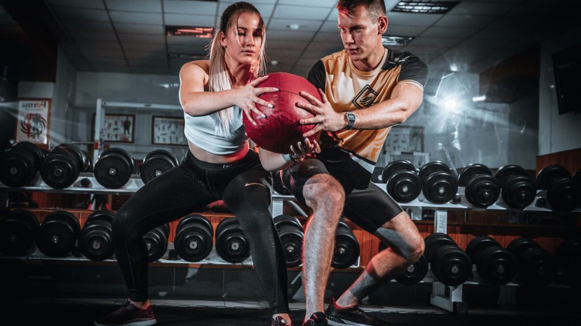 A man and woman perform a tandem workout with a medicine ball in a gym setting.