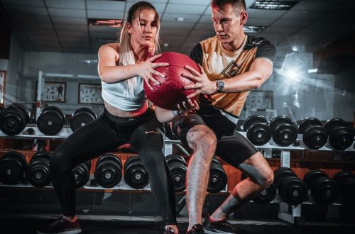 A man and woman perform a tandem workout with a medicine ball in a gym setting.