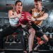 A man and woman perform a tandem workout with a medicine ball in a gym setting.